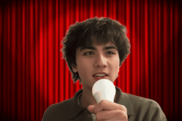 Spotlight on young male with dark curly hair holding a microphone. Red theatre curtain background.