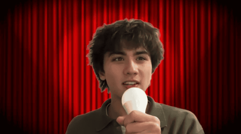 Spotlight on young male with dark curly hair holding a microphone. Red theatre curtain background.