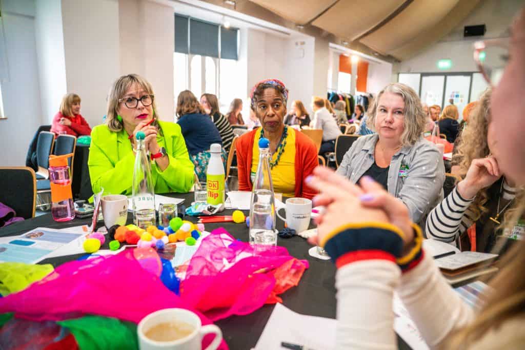 group of 3 women sat round a table listening carefully. The table is full of bright coloured fabric and pompoms