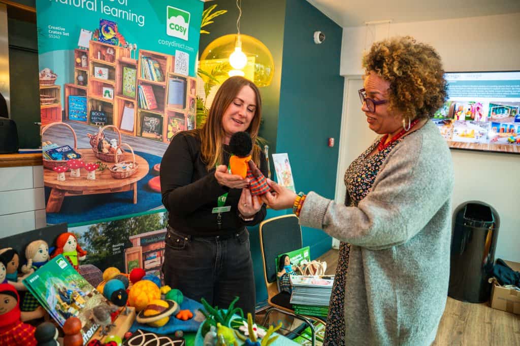 2 women stood by a stall. One woman is showing the other woman a child's toy