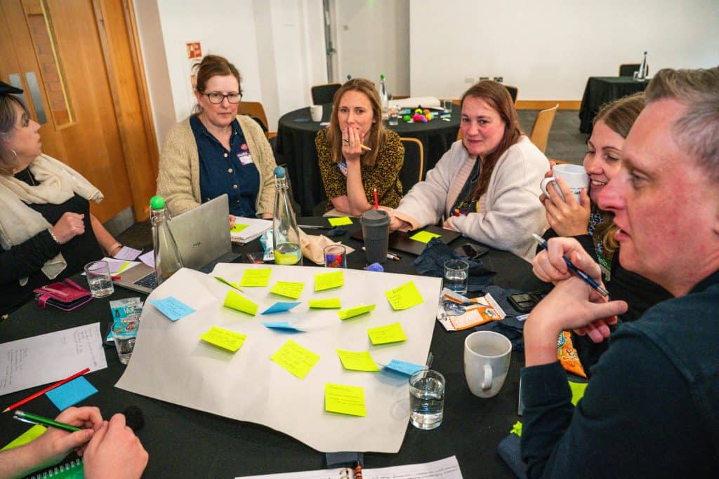 group of 5 women and 1 man sat round a table having a discussion. The table contains large sheet of paper with post-it notes
